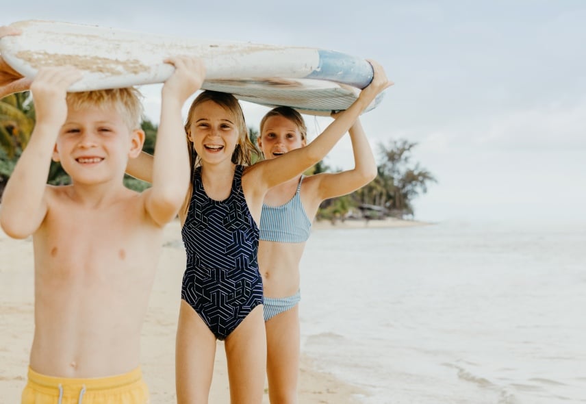 Kids playing with a surfboard on a sunny beach while parents relax - Heritage Resorts